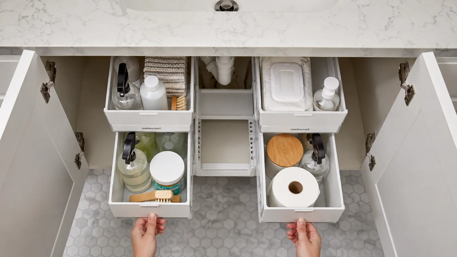 Hand pulling out the lower sliding drawer of a white 2-tier under-sink organizer stocked with dish soap and scrubbing brushes